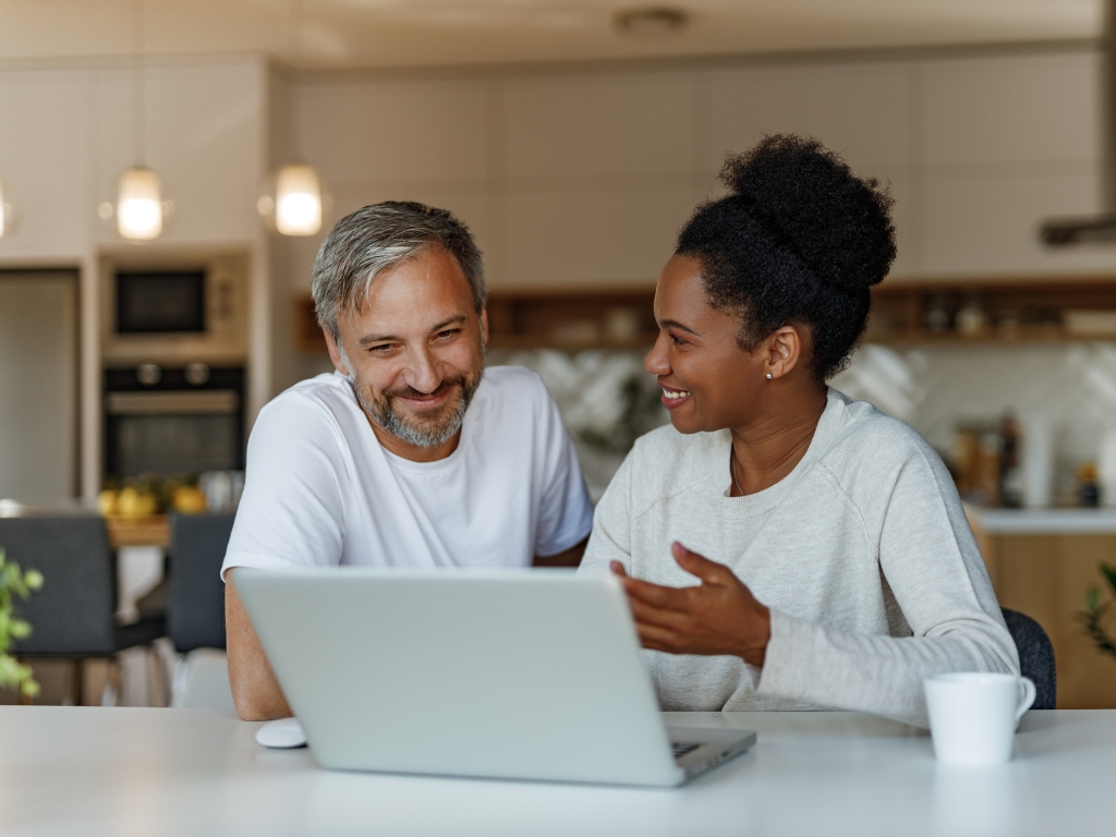 Alt text: Two people sitting at a table in a kitchen with an open laptop and a coffee cup, engaged in conversation.